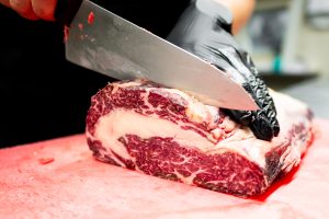 Gloved hand slicing raw steak on a red cutting board in a butcher or meat processing facility, illustrating UV decontamination solutions for knives, conveyor belts and air.
